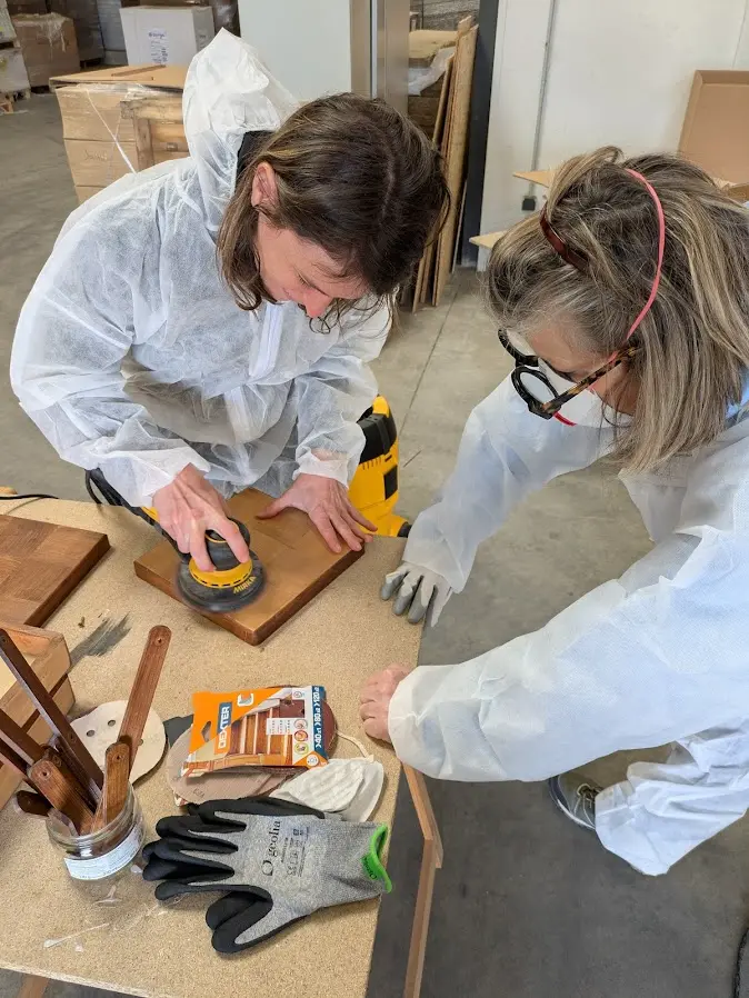 Deux femmes en tenue de protection travaillent ensemble sur un projet de menuiserie, utilisant des outils pour façonner des matériaux en bois sur une table.