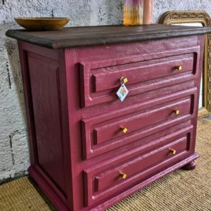Commode Adelphe – Commode en bois bordeaux avec trois tiroirs ornés de boutons dorés, surmontée d'un bol simple, placée devant un mur texturé.