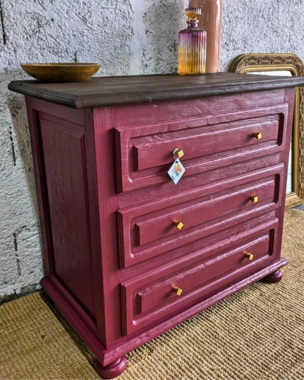 Commode Adelphe – Commode en bois bordeaux avec trois tiroirs ornés de boutons dorés, surmontée d'un bol simple, placée devant un mur texturé.