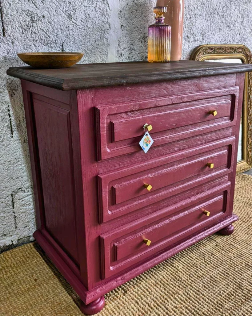 Commode Adelphe – Commode en bois bordeaux avec trois tiroirs ornés de boutons dorés, surmontée d'un bol simple, placée devant un mur texturé.