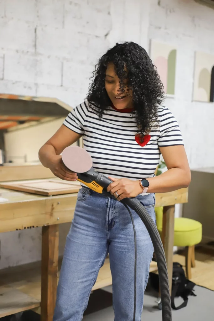 Mary tenant une ponceuse électrique, prête à travailler, vêtue d'une chemise rayée avec un cœur rouge et d'un jean bleu dans son atelier.