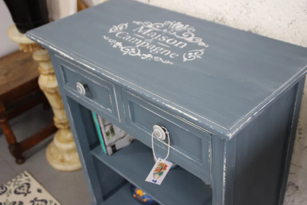 Meuble de Rangement – Armoire vintage bleue avec un design décoratif sur le dessus, comprenant un tiroir et une étagère ouverte, ornée d'étiquettes.