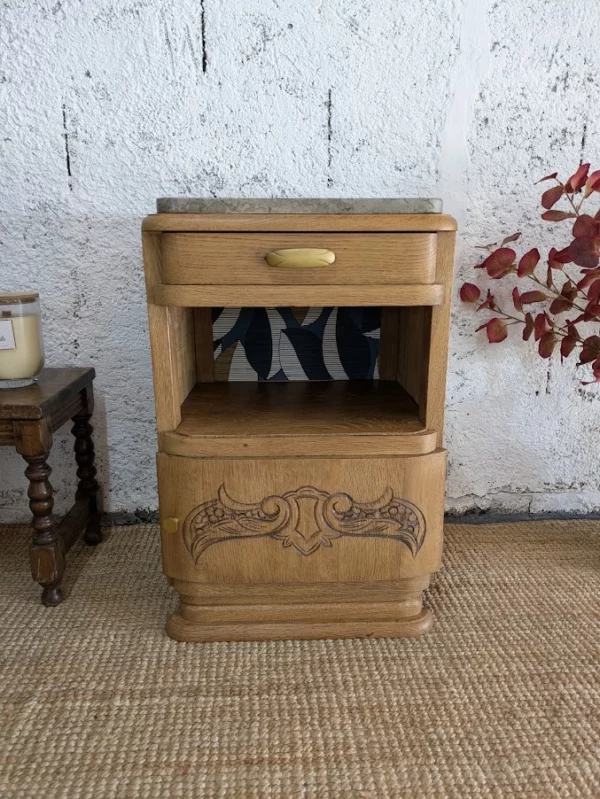 Table de chevet Florent – Table de chevet vintage en bois avec des sculptures complexes, un tiroir et une étagère ouverte, placée devant un mur texturé et un tapis tissé.