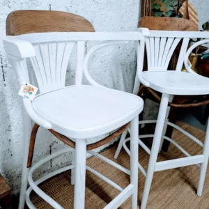 Paire de chaises de Bistrot – Deux tabourets de bar élégants avec des cadres en bois blanc et des assises en rotin tressé, placés devant un mur texturé.