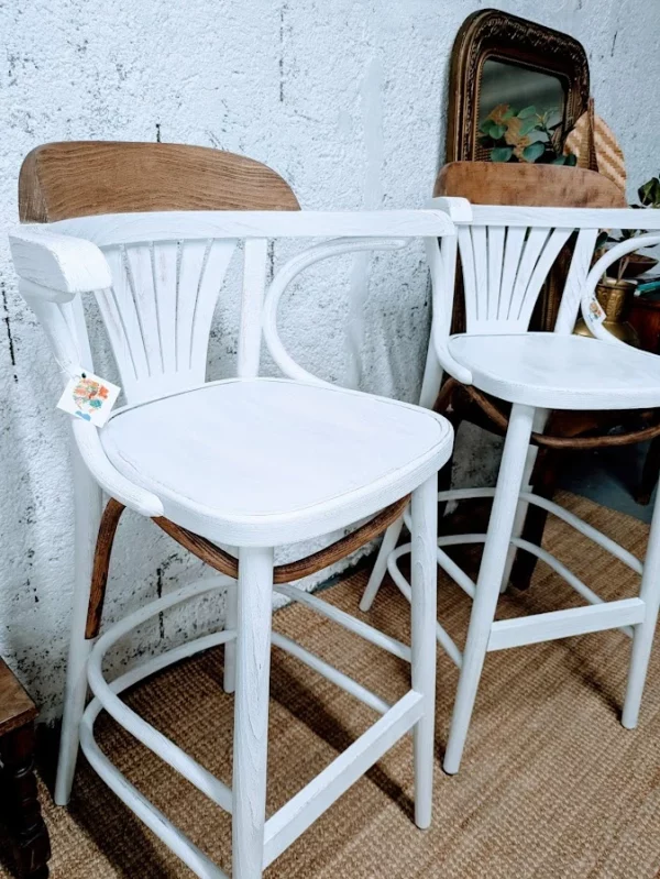 Paire de chaises de Bistrot – Deux tabourets de bar élégants avec des cadres en bois blanc et des assises en rotin tressé, placés devant un mur texturé.